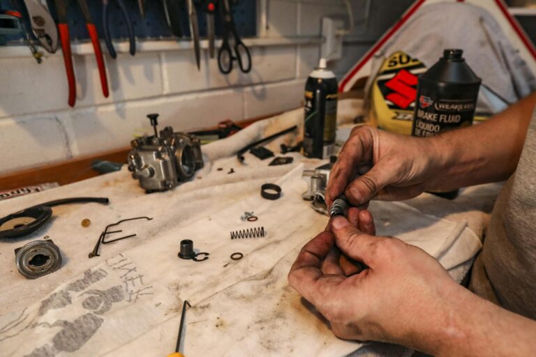 Close-up of hands repairing a carburetor with tools on a workbench in a garage setting.